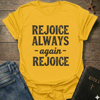 Yellow t-shirt with 'REJOICE ALWAYS again REJOICE' text Philippians 4:4 bold faith apparel Faith Forward on a wooden surface with jeans and a Bible.