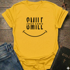 A yellow short-sleeve t-shirt with a black graphic reading 'SMILE JESUS CARES SMILL' and a smiley face, styled flat on a rustic wooden surface with blue jeans and a Bible nearby.