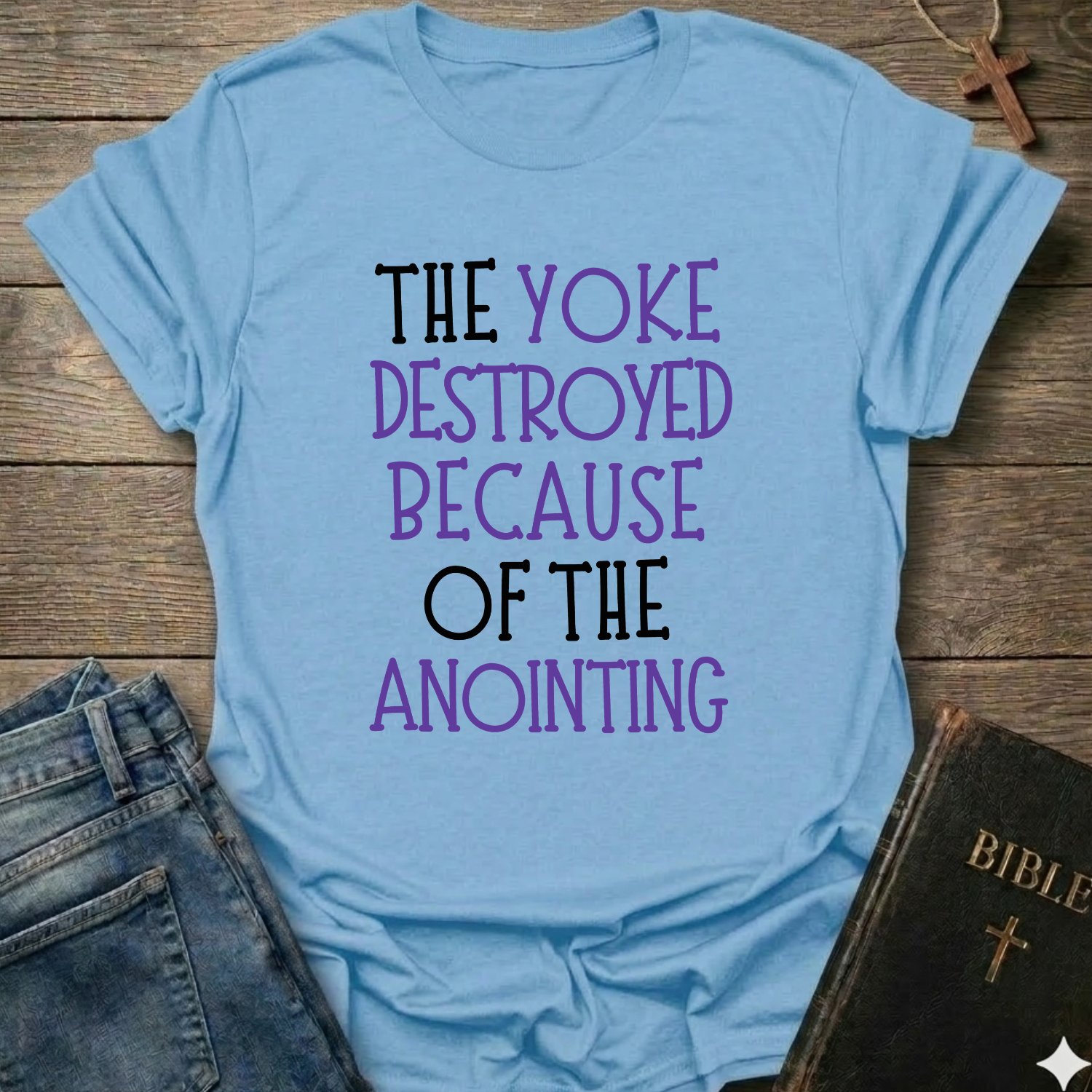 Light blue t-shirt with motivational text 'The yoke destroyed because of the anointing' Isaiah 10:27 faith apparel conversation starter on a wooden surface with jeans and a Bible.