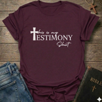 Maroon t-shirt with 'This is my testimony shirt' text and cross minimal faith apparel conversation starter. It is on a wooden surface with jeans and a Bible.